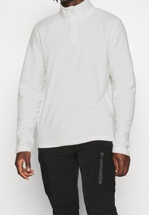 Homme portant un pull à fermeture éclair blanche au quart et un pantalon cargo noir avec des poches zippées, debout devant un fond uni.