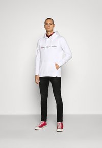 Balta medvilninė su kapuce megztinė su "DENIM PROJECT" spauda, kengūros kišenė ir raišteliai. Sujungta su juodomis džinsomis ir raudonais sportbačiais.