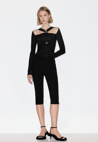 Vrouw gekleed in een zwarte nauwsluitende outfit met uitsneden op de schouders, cropped broek en zwarte hoge hakken met pluche bandjes, staand tegen een witte achtergrond.