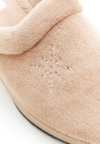 Chausson beige avec une surface douce et texturée, doté d'une sangle arrondie et de strass décoratifs disposés en motif étoile. Semelle noire.