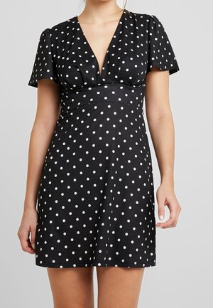 Kobieta ubrana w czarną sukienkę z krótkim rękawem w białe groszki z głębokim dekoltem w kształcie litery V, stoi na tle jednolitego szarego tła.