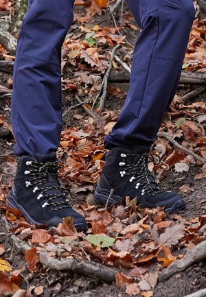Schwarze Wanderschuhe mit grauen und gelben Schnürsenkeln, ausgestattet mit einem Obermaterial aus Wildleder und synthetischen Materialien, robust gestaltet für unwegsames Gelände, auf einem mit Blättern bedeckten Boden.