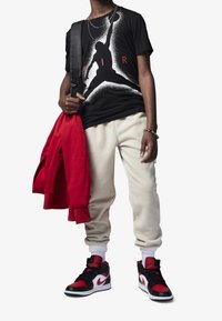 Czarna koszulka z białą grafiką i czerwoną napisami, beżowe dresy i czarno-czerwone sneakersy. Czerwona bluza zarzucona na ramię, srebrna bransoleta.