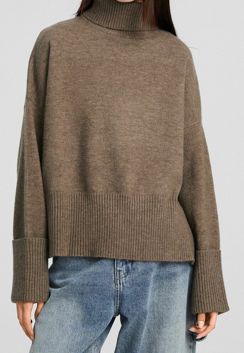 Femme portant un pull à col roulé côtelé marron avec des poignets prolongés et un jean bleu délavé, debout devant un fond uni.