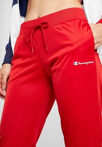 Pantalon de sport rouge avec taille à cordon et logo Champion sur la cuisse droite porté par une personne avec la main dans la poche et le ventre visible.