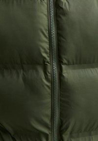 Giubbotto imbottito verde oliva con zip verticale. Presenta una texture trapuntata e una finitura liscia e opaca. La zip è nera con un tirante arrotondato.