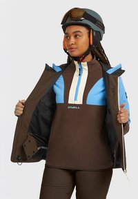 Brūns, zils un krēmkrāsas flīsa džemperis ar puszipa apkakli. Kombinēts ar brūnu jaku, ķiveri un sniega brillēm.