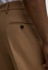 Brązowe wełniane spodnie z tylną kieszenią ozdobioną czarnym guzikiem, teksturowana tkanina oraz dopasowany krój widoczny z boku.