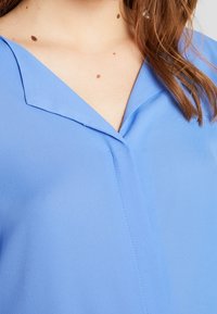 Světle modrá blůza s výstřihem do V, měkkou texturou látky a jemným švem podél přední strany. Doplněná límcem.