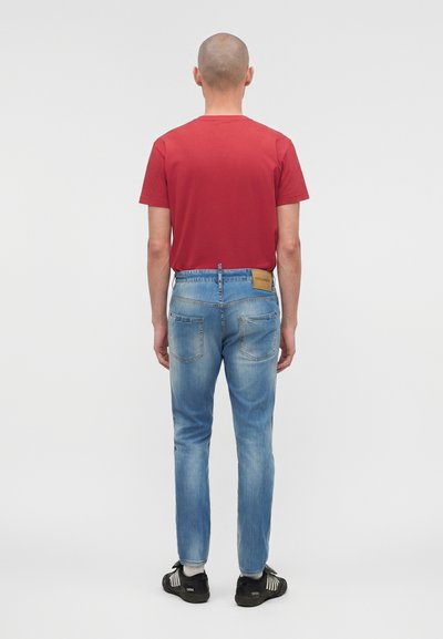 Άνδρας που στέκεται με την πλάτη προς τα εμπρός, φορά κόκκινο μπλουζάκι, ανοιχτόγαλαντζιά τζιν με δερμάτινο ταμπελάκι, λευκές κάλτσες και μαύρα αθλητικά παπούτσια.