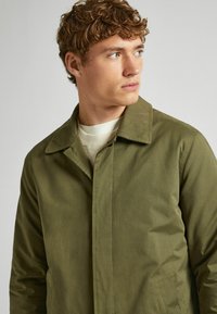 Olijfgroene jas met een kraag en knoopsluiting, met een gladde textuur en twee zijzakken. Gdragen over een crèmekleurige trui.