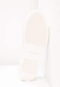 Semelle de chaussure blanche avec motif ondulé et logo "ALDO" embossé au centre, positionnée droite contre un coin blanc.