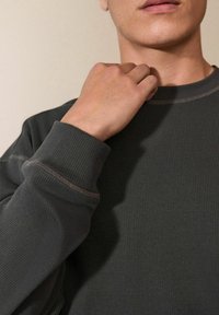 Grijze geribbelde sweatshirt met een klassieke ronde hals en gestructureerde stof. Kenmerkt zich door contrasterende stiksels en aansluitende manchetten. Beeld van een halve lichaamsweergave van een hand die de kraag aanpast.