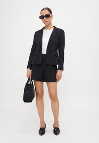 Svart skräddarsydd kavaj med fickor, kombinerad med svarta shorts. Vit topp under, svarta fyrkantiga solglasögon och en svart axelväska. Glänsande svarta skor.