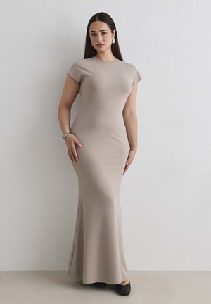 Femme aux longs cheveux foncés portant une robe maxi beige ajustée, des chaussures noires, une montre argentée et des boucles d'oreilles blanches, debout contre un mur uni.