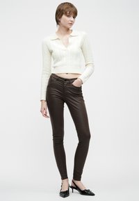 Mladá žena stojí s rukou v vrecku, oblečená v krémovom krátkom svetrí s pleteným vzorom, tmavých slim fit nohaviciach a čiernych lodičkách na podpätku.