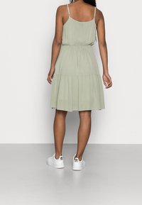 Ljusgrön, ärmlös klänning med ett lager-på-lager-design och resår i midjan. Modellen bär vita sneakers. Slät tyg, avslappnad passform.