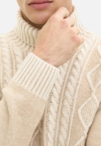 Beżowy sweter z dzianiny w warkocze z wysokim, prążkowanym kołnierzem oraz różnymi wzorami teksturowymi na korpusie i rękawach. Częściowo widoczna ręka.