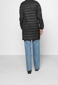 Cappotto trapuntato nero con maniche lunghe, che raggiunge la metà della coscia. Presenta cuciture orizzontali e un colletto alto, abbinato a jeans azzurri chiari.