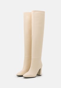 Botas de imitación de cuero crema hasta la rodilla, con punta cuadrada, tacón grueso y textura suave. Presentan una silueta ajustada con costuras mínimas.