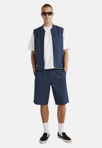 Chaleco sin mangas azul marino sobre una camiseta blanca, combinado con shorts azul marino. Los accesorios incluyen gafas de sol oscuras y zapatos negros sin cordones.