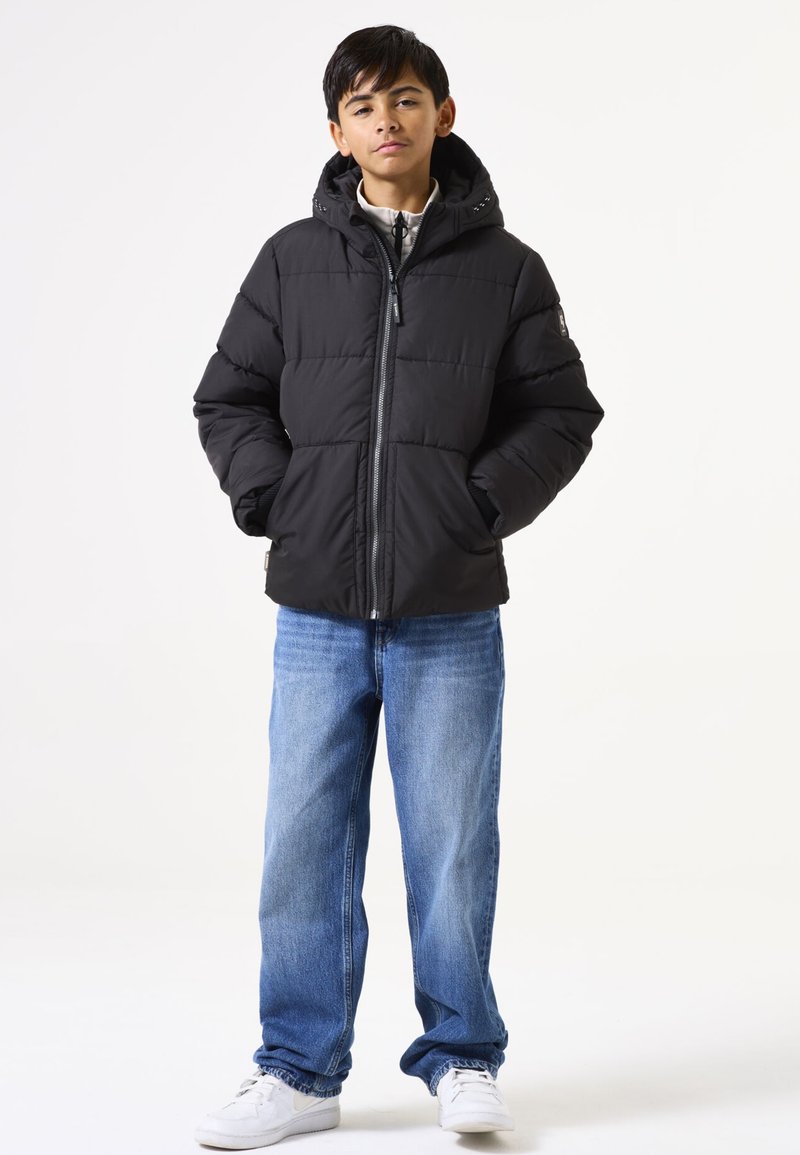 Chaqueta de plumas negra con capucha, cierre de cremallera y paneles cosidos. Combinada con vaqueros azules y zapatillas blancas en un modelo de pie.