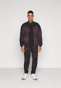 Melna un tumši sarkana gradienta treniņtērps ar rāvējslēdzēja jaku un elastīgām aprocēm, kuram ir neliels zaļš logo uz krūtīm un biksēm.