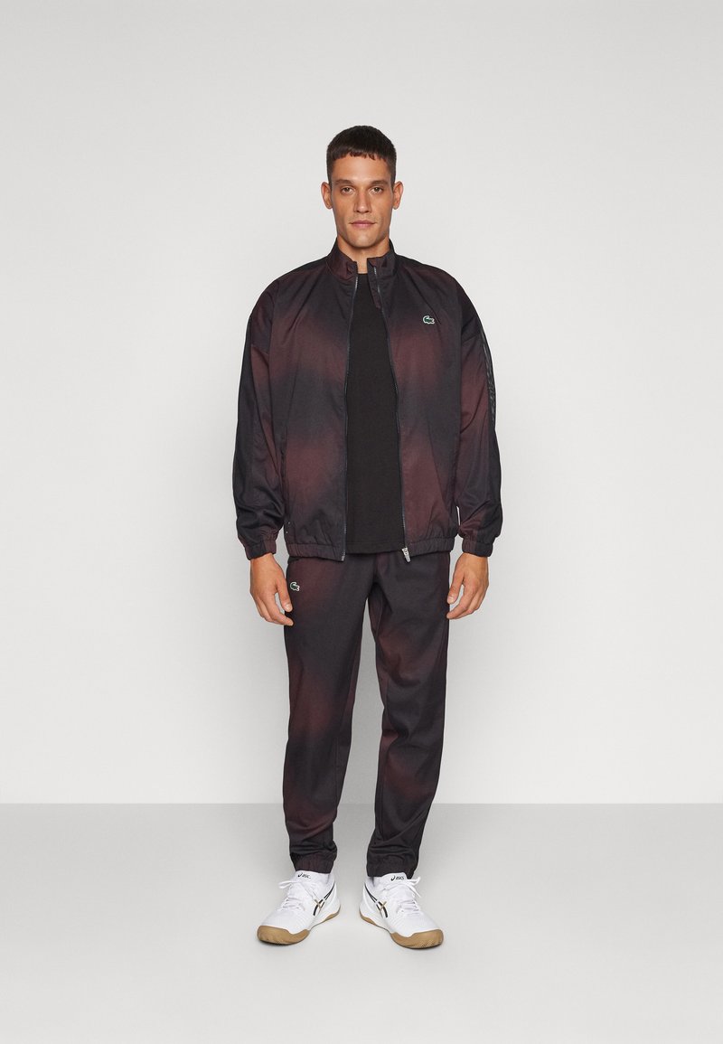 Schwarz und dunkelroter Farbverlauf Trainingsanzug mit einer Jacke mit Reißverschluss und elastischen Bündchen, ausgestattet mit einem kleinen grünen Logo auf der Brust und an der Hose.