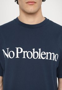 Námořnická modrá bavlněná tričko s bílým grafickým potiskem s textem "No Problemo" v tučném, moderním písmu. Krátké rukávy a kulatý výstřih.