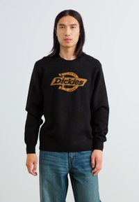 Schwarzer Pullover mit einem braunen "Dickies"-Logo in der Mitte, langen Ärmeln, Rundhalsausschnitt und weicher Textur. Kombiniert mit blauen Jeans.