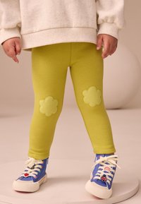 Grüne Leggings mit blumenförmigen Kniepolstern, kombiniert mit blauen Schuhen mit bunten Akzenten und weißen Schnürsenkeln, getragen zu einem leichten Pullover.