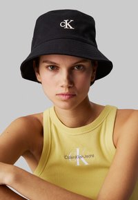 Schwarzer Bucket Hat aus strapazierfähigem Stoff, mit einem weißen, gestickten "CK"-Logo auf der Vorderseite. Getragen mit einem gelben, gerippten Tanktop.