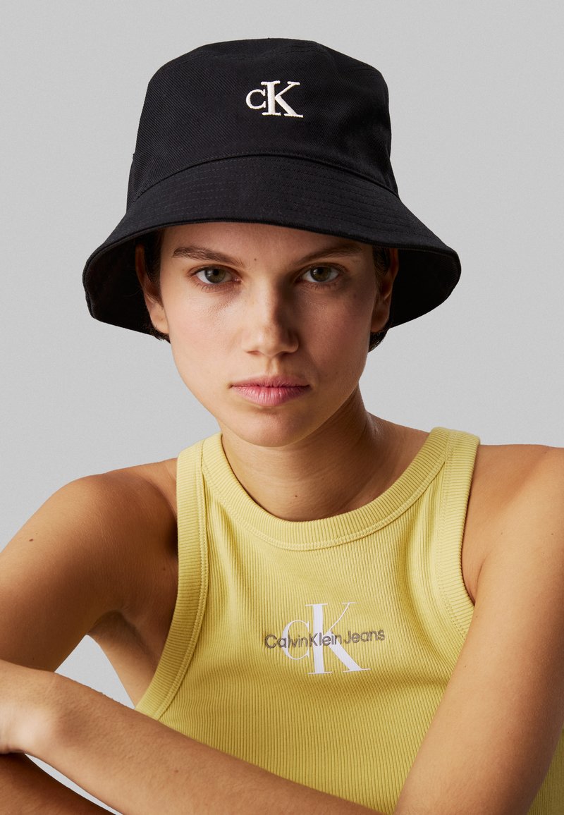 Schwarzer Bucket Hat aus strapazierfähigem Stoff, mit einem weißen, gestickten "CK"-Logo auf der Vorderseite. Getragen mit einem gelben, gerippten Tanktop.