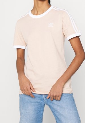 Personne portant un t-shirt beige Adidas avec bordure blanche et rayures sur les manches, associé à un jean bleu clair, seul le torse est visible.