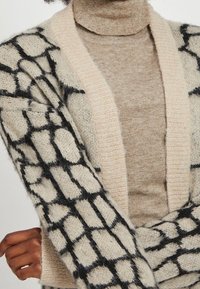 Mönstrad cardigan i krämfärgat med svarta geometriska designer, med en mjuk textur, öppen front och långa ärmar, lagd över en beige polotröja.
