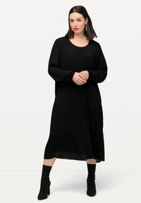 Musta pleatattu midi-mekko, jossa on pitkät hihat, pyöreä pääntie ja elastiset hihansuut, yhdistettynä mustiin nilkkureihin. Sileä pinta, minimalistinen muotoilu.