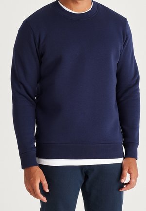 Man draagt een effen marineblauwe sweatshirt met lange mouwen over een wit overhemd, gecombineerd met donkerblauwe broek, staat met ontspannen armen.