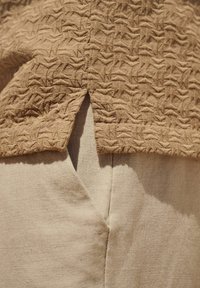 Haut texturé tan présentant un motif, avec une fente à l'ourlet, associé à un tissu beige clair, créant un look superposé.