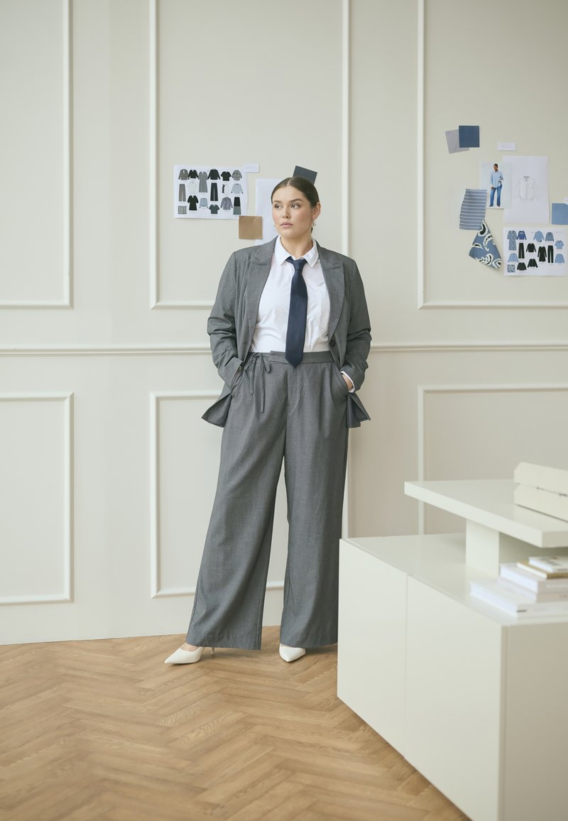 Costume gris avec une veste ajustée et un pantalon à jambes larges, accompagné d'une chemise blanche et d'une cravate foncée. Des chaussures blanches à bout pointu complètent le look.