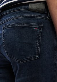 Tmavomodré rifle z denimu so stredným vypraním, s koženou značkou s vyrazeným textom a malým trojfarebným logom na zadnom vrecku.