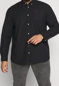 Svart skjorta med knäppning framtill och bröstficka, samt vita knappar. Tillverkad av ett slätt tyg, utrustad med spetsig krage och långa ärmar.