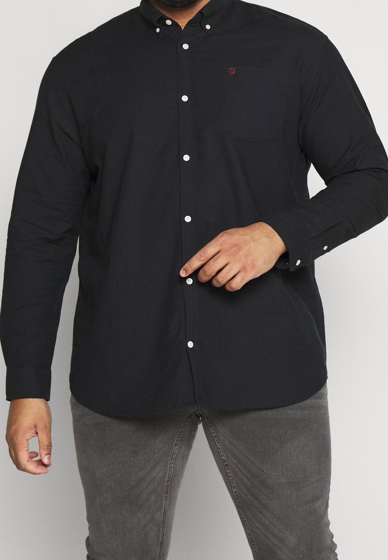 Svart skjorta med knäppning framtill och bröstficka, samt vita knappar. Tillverkad av ett slätt tyg, utrustad med spetsig krage och långa ärmar.