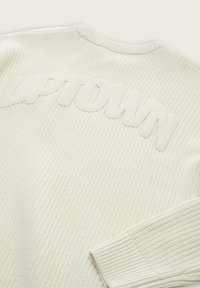 Pull en tricot blanc avec le lettrage en relief "UPTOWN" dans le dos, présentant des poignets côtelés et une texture dans tout le tissu.
