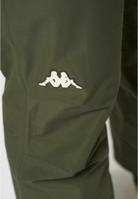 Olívazöld nadrág könnyű anyagból, szemrevaló fehér logóval a bal combon, sima textúrával és testhezálló szabással.