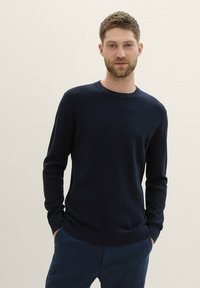 Marineblauwe gebreide trui met een ribbelstructuur, ronde hals en lange mouwen, gecombineerd met marineblauwe broeken. Eenvoudig ontwerp, zonder patronen of accenten.