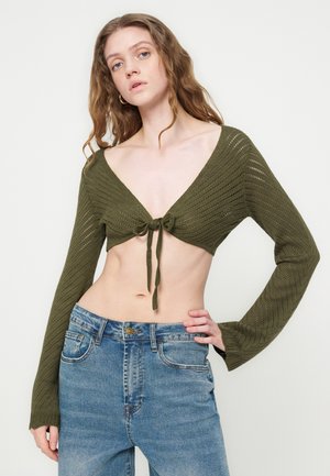 Femme aux longs cheveux ondulés portant un crop top en maille vert olive avec un nœud à l'avant et un jean bleu taille haute, debout devant un fond blanc.