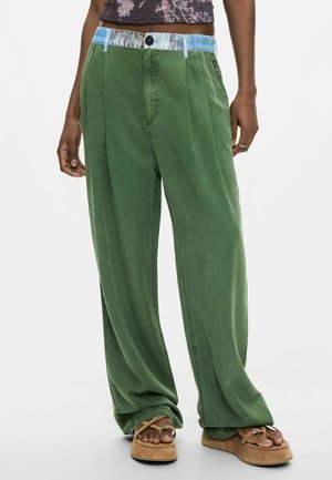 Persona che indossa pantaloni verdi larghi con coulisse in vita e sandali marroni, in piedi davanti a uno sfondo bianco neutro.