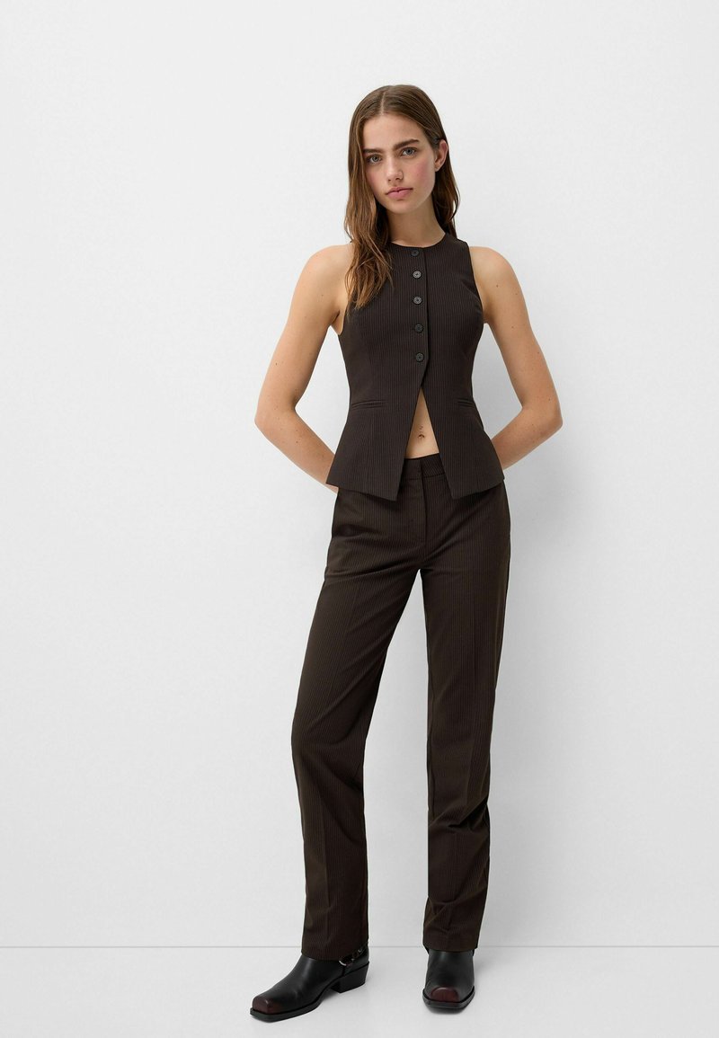 Canotta marrone su misura con scollatura alta e cinque bottoni frontali, abbinata a pantaloni slim-fit coordinati. Tessuto liscio con una leggera texture.