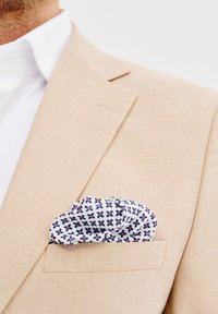 Beżowa marynarka ze strukturalnym rewersem, z białą poszetką z powtarzającym się niebieskim i czerwonym geometrycznym wzorem.