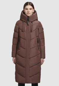 Kvindelig iført en lang, brun quilted pufferjakke med høj krave, justerbar hætte og lynlåslommer på siden, stående foran en ensfarvet baggrund.